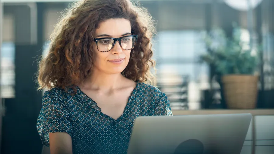 A woman wearing glasses with curly brown hair is sitting and working on her laptop.