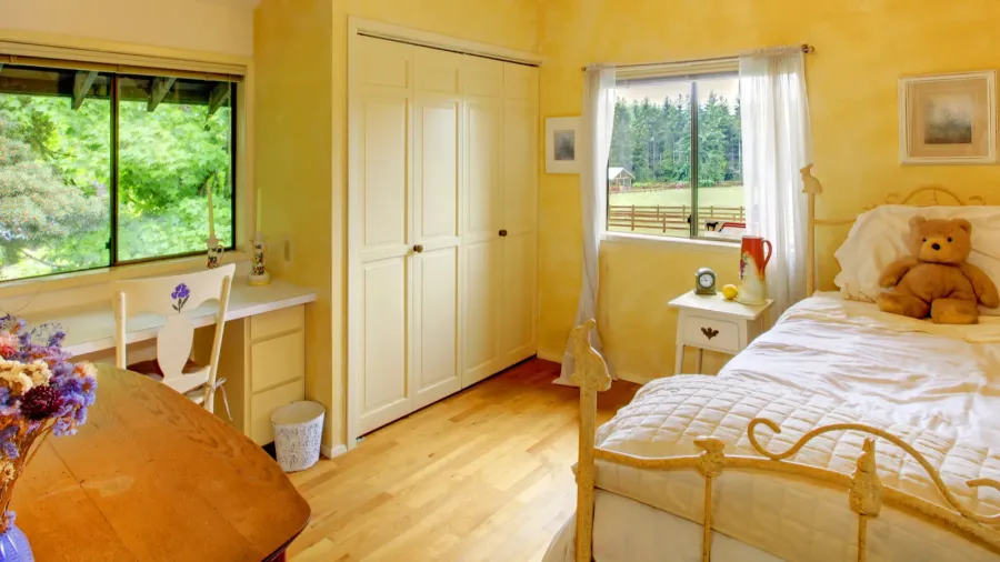 A bright yellow room with a bed, teddy bear, and a window looking out to greenery.