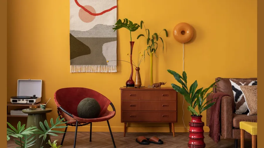 A cozy room with a bright yellow wall, featuring a red chair, a wooden sideboard, plants, and a colorful wall hanging.