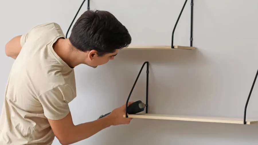 A young man installing floating shelves on a white wall.