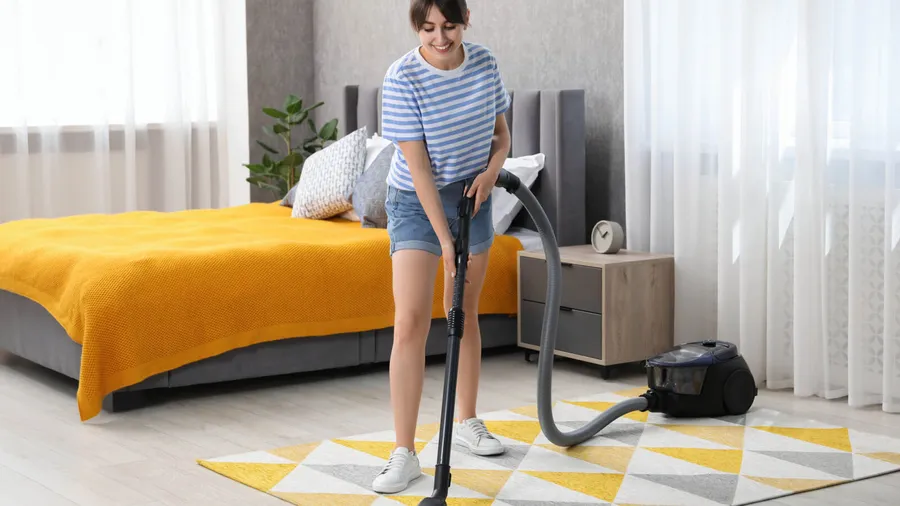 A woman vaccumming in a bedroom with cozy gray bed, nightstand with wheels, and a geometric patterned rug.