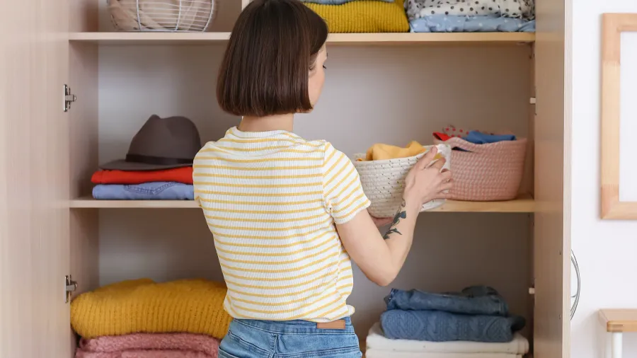 A woman organizing clothes on a shelf while holding a woven basket.
