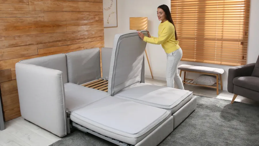A woman is assembling a modern convertible sofa, adjusting the fabric sections to create a flat sleeping surface in a bright and stylish living room.