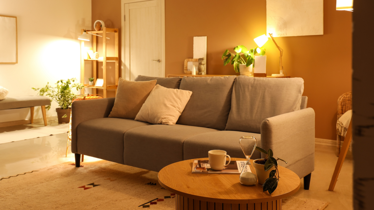 A cozy living room with a gray sofa, a wooden coffee table, and warm lighting. There are plants, pillows, and a mug on the table.