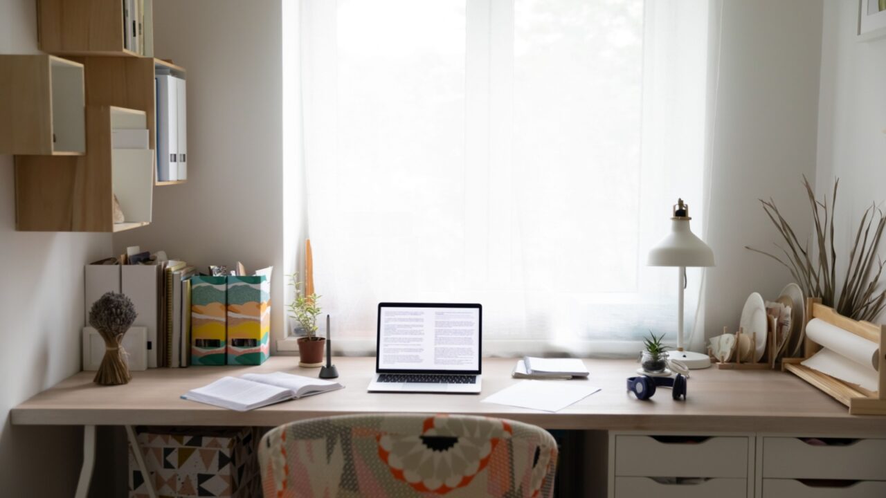 A study table with laptop, lamp and book against a window with white curtains and wooden floating shelves on the adjacent wall.