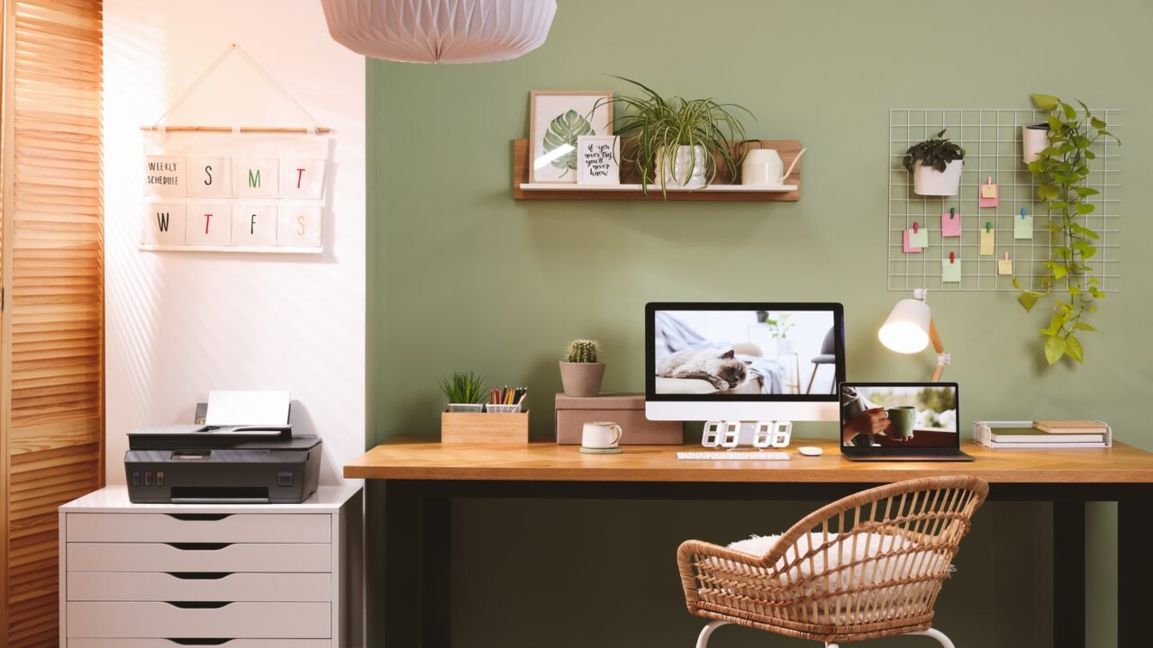 A home office with a wooden desk, a woven chair, and a green wall. There is a computer, a laptop, a printer, and various plants and decorations in the room.