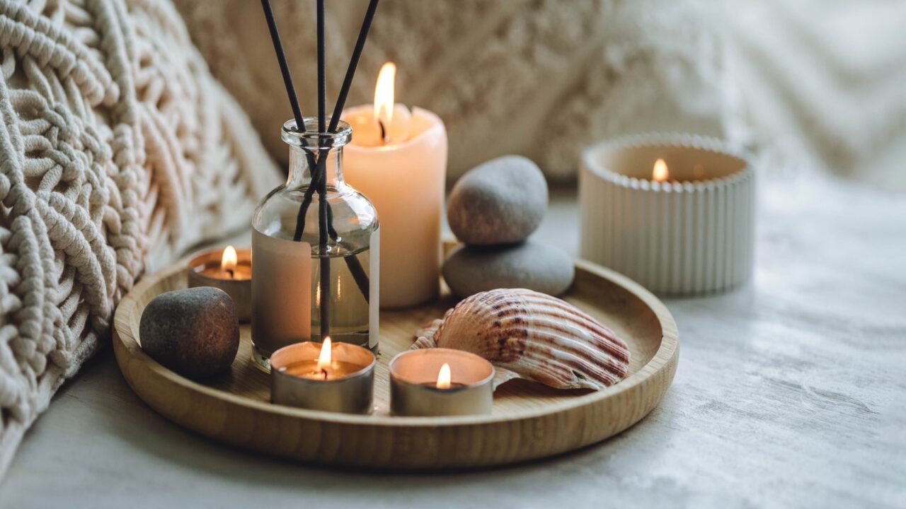 An essential oil reed diffuser and candles on a bamboo tray placed on a bed.