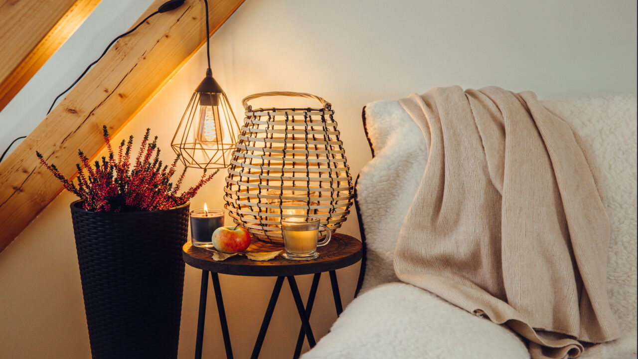 A sheepskin cover on a sofa with a wooden table beside it with woven lantern and candles on it.