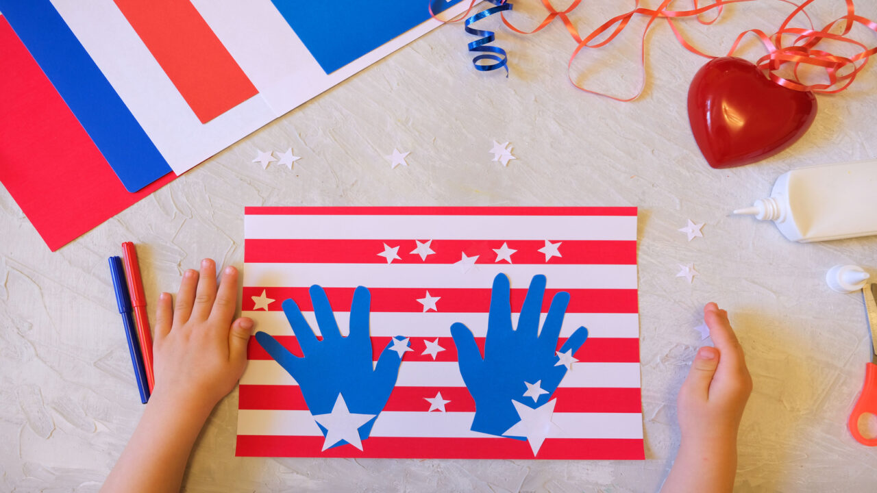 A kid making USA flag themed DIY card using red, white, and blue papers.