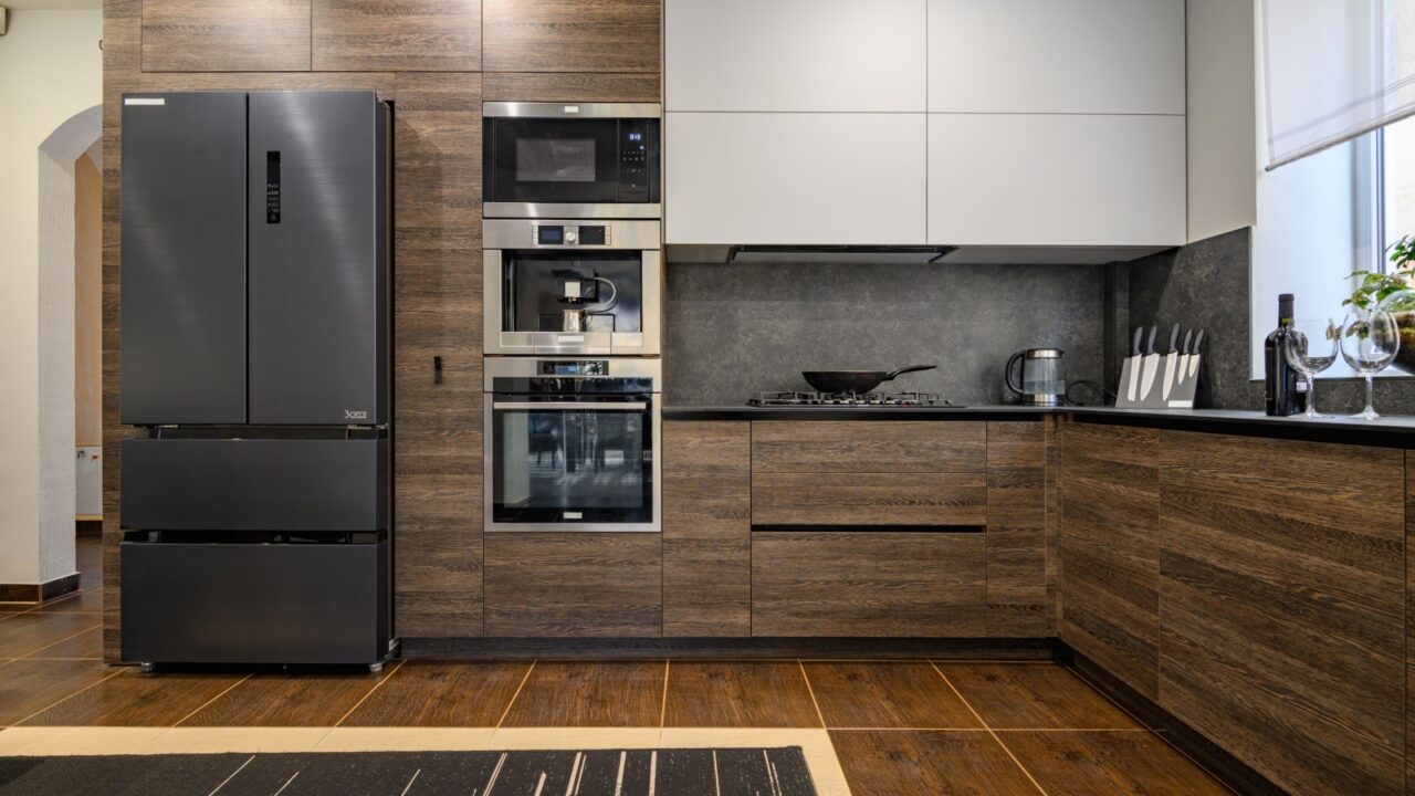 A modern kitchen with dark wood cabinets, stainless steel appliances, and a gray countertop. There is a refrigerator, microwave, coffee maker, oven, stovetop, and a knife block.