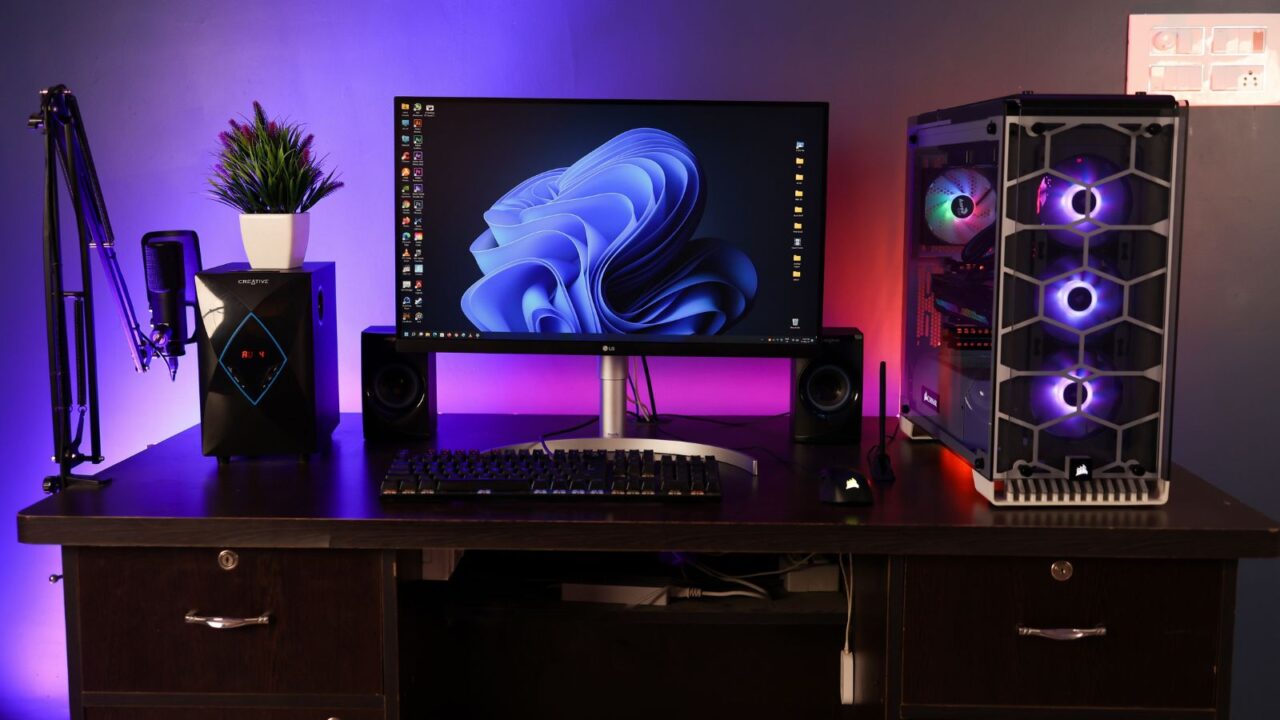 A high-end gaming setup with desktop, CPU, speakers, and mic on a wooden table in front of a dark background and RGB lights.