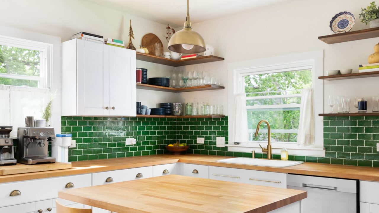 A kitchen interior with butcher block wood countertops, white cabinets, a gold fixture hanging over the island, and green subway tile backsplash.