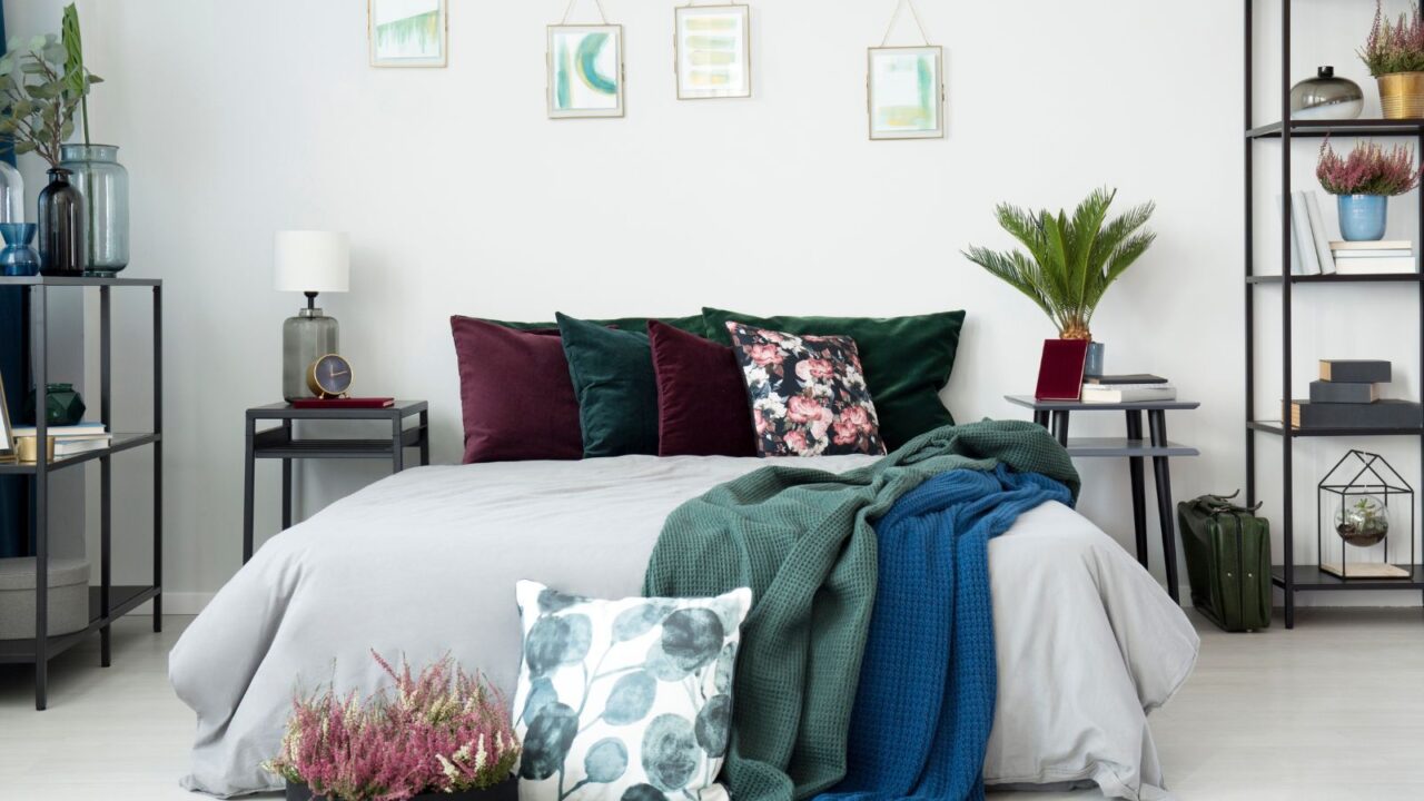 A stylish bedroom interior with comfortable bed with green and purple cushions, blankets, plants, small posters and bedside tables.