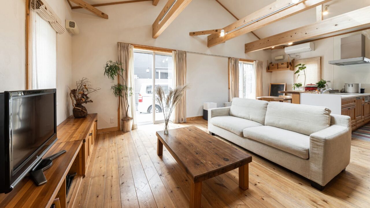 A stylish rustic living room interior with sofa, wooden table, built-in wooden cabinets with TV, houseplants, hardwood flooring, and an open kitchen.