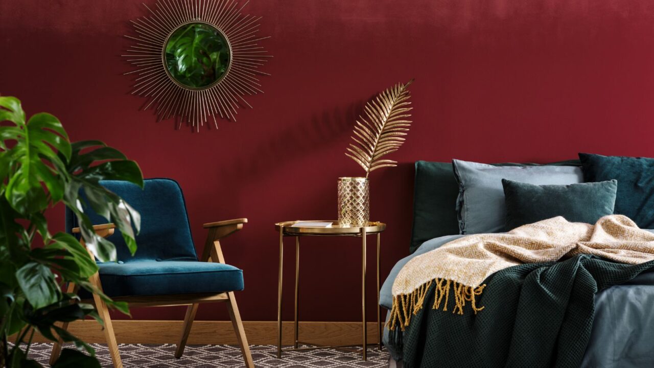 Gold table between green armchair and bed in sophisticated red bedroom interior with mirror