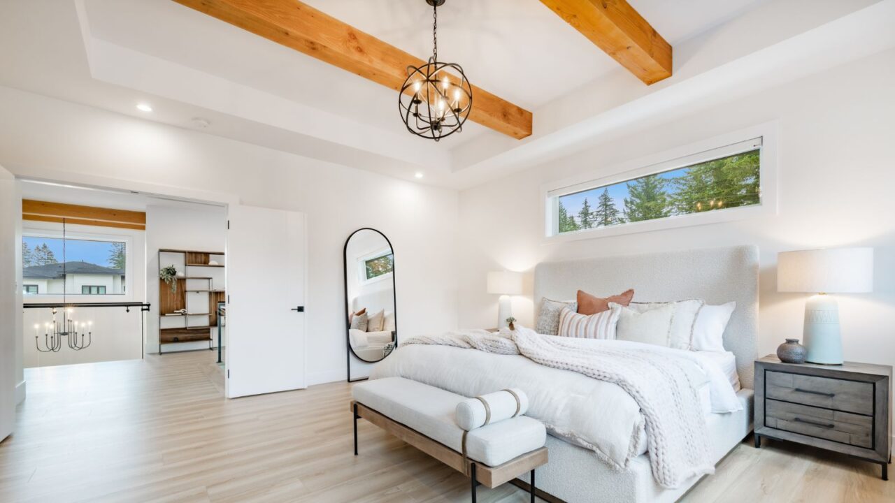 Primary master bedroom with wood beam ceiling, hardwood floors, white walls, a metallic chandelier, and oval mirror.