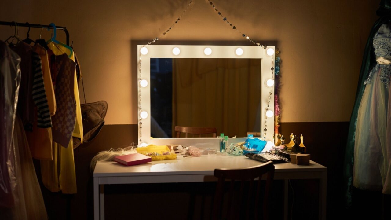 Vanity mirror placed on a white table in dressing room. Racks of clothes are placed in the room too.