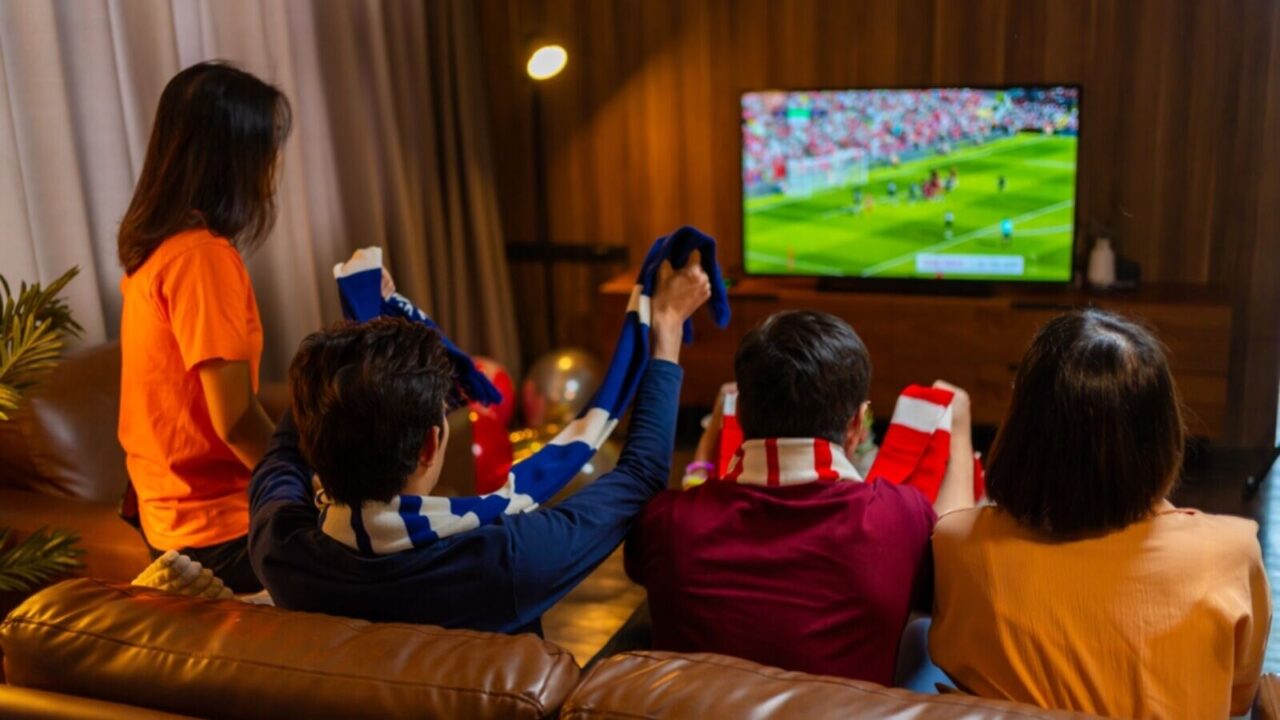 Group of friends sitting on sofa watching and cheering football or soccer games competition on TV together at home.