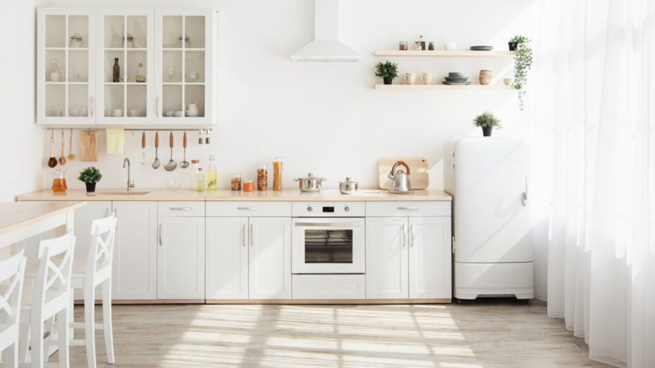 Kitchen with light walls, white furniture and shelves with crockery and plants in pots, small refrigerator in dining room.