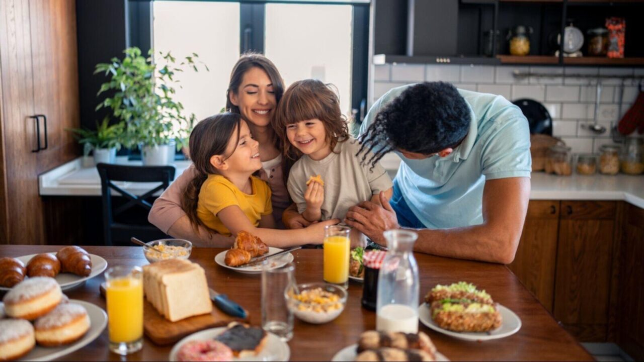 Happy parents having breakfast at kitchen table with children at home, looking at them with love and kindness.