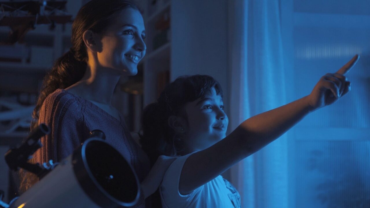 A mother and daughter looking at stars through the window. A telescope is also placed beside them