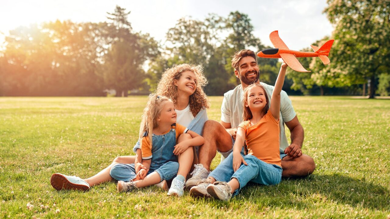 Happy young family dad, mom and two children daughters playing with an airplane sitting in a meadow on the grass in the backyard