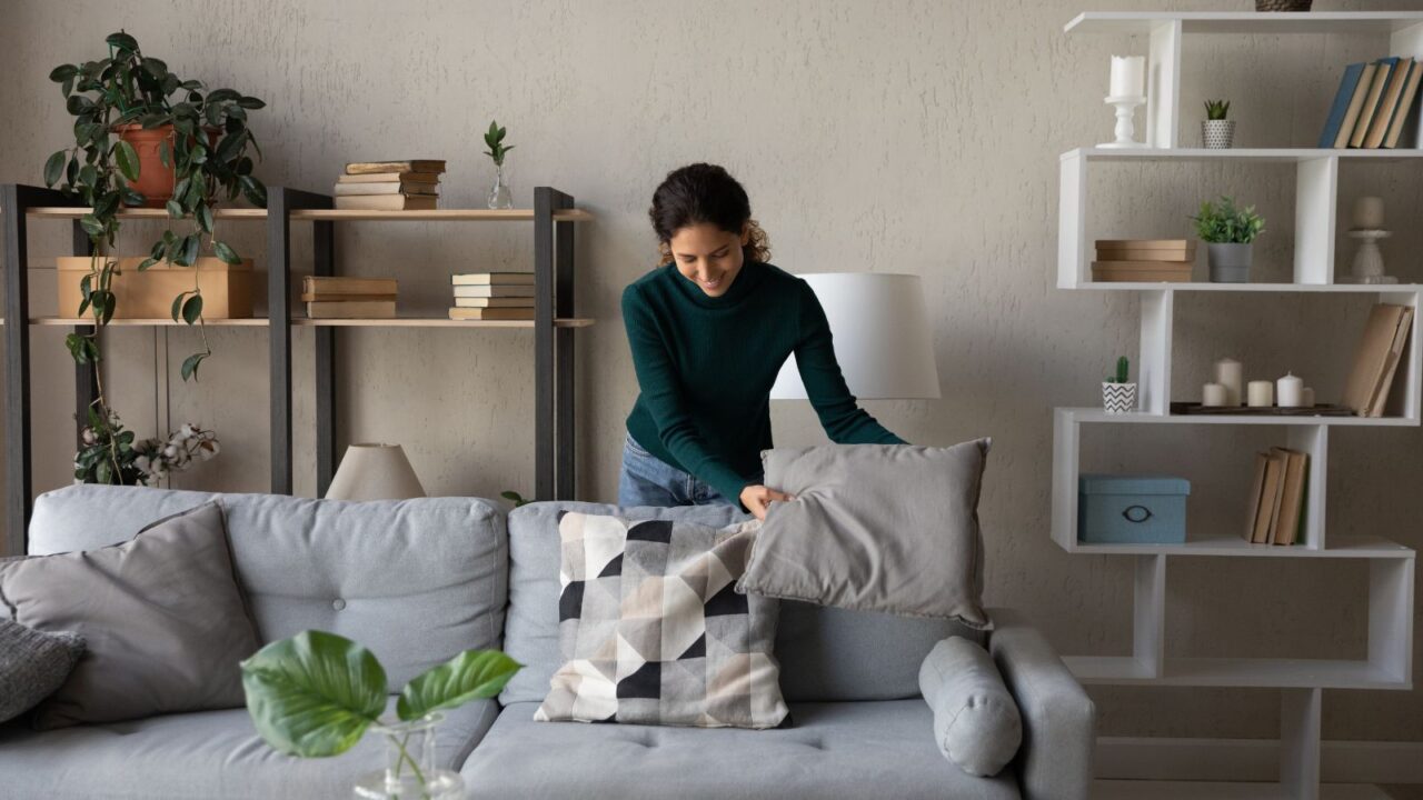 A woman arranging pillows on a gray sofa in a living room with bookshelves and plants in the background.