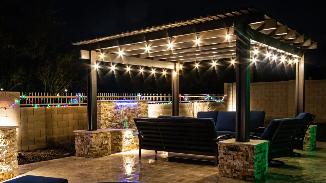 A resort style backyard at night with a waterfall, pergola, and a firepit at night.