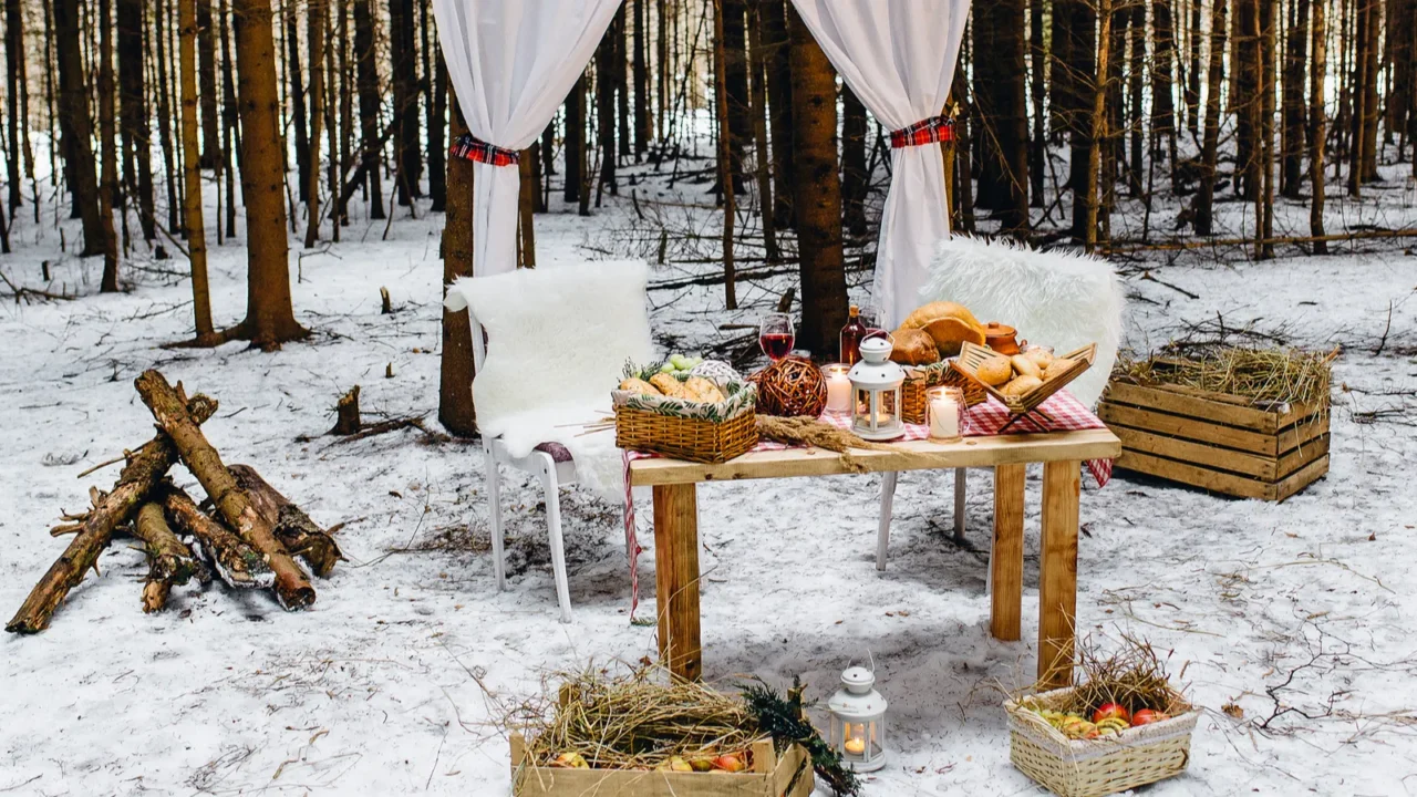 Cornice with white curtains , wooden crates with hay, pine wood, white fur covered chairs, a table with bread and romantic candles. Picnic in the winter