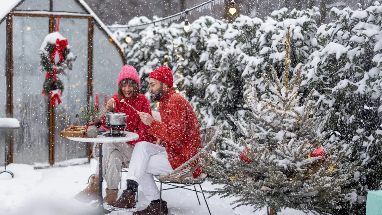 Man and woman have romantic dinner with fondue, while sitting together by the table at beautifully decorated snowy backyard. Young family celebrating winter holidays outdoors