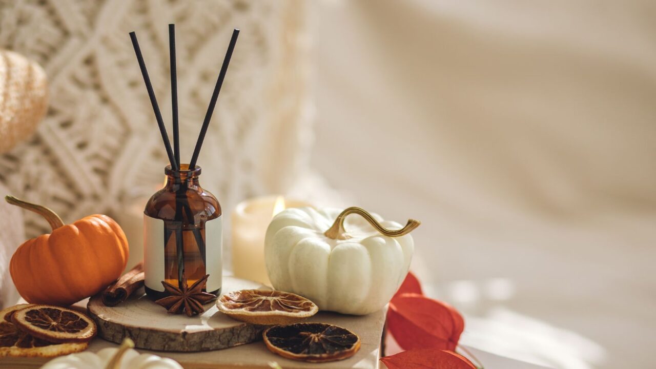 A reed diffuser with fall decor including leaves and pumpkin pie are on a book. Cinnamon sticks, cloves, and dried citrus are also present. Cozy fall knitted wool fabric is also in the background