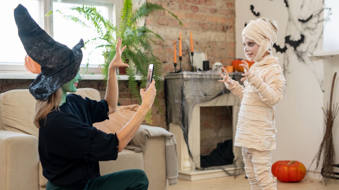 Female in witch costume taking photo of a kid in zombie attire in living room.