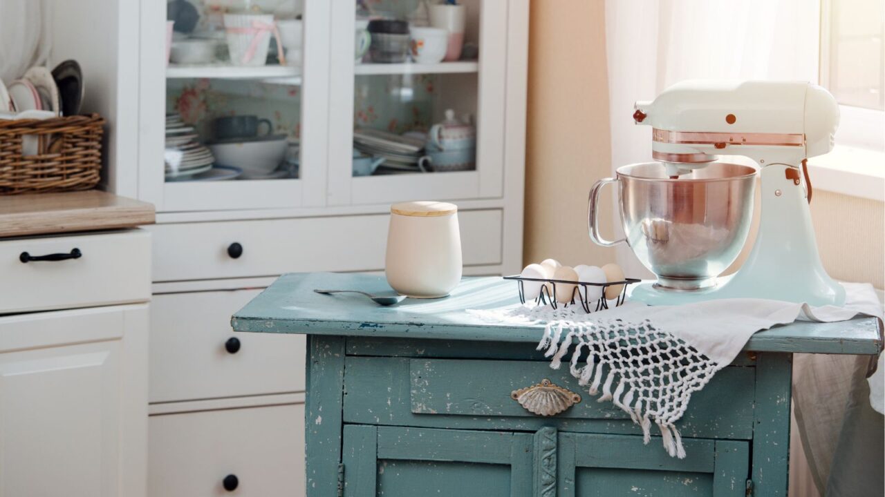 Baking tools on a green table inside the kitchen. Mixer, eggs, jar, and spoon in the baking corner.