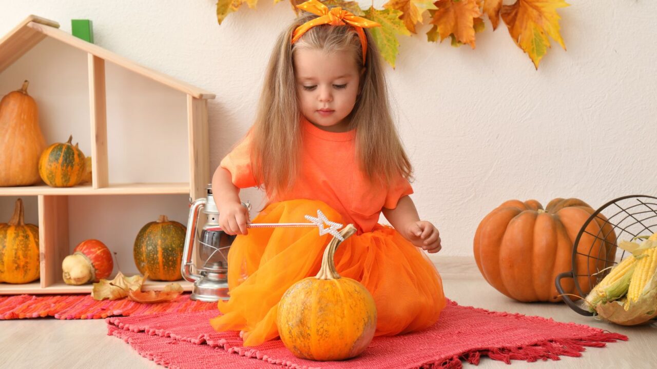 Little girl sitting on a red rug in a playroom with magic wand and pumpkin. Pumpkins are lying on the floor and fall leaves are hanging on the wall. The girl is dressed up in orange frock and band according to the fall season.