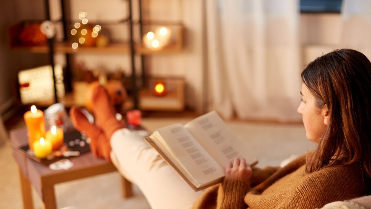 young woman is reading a book on a sofa in a cozy ambiance and many candles are placed on the coffee table and shelf