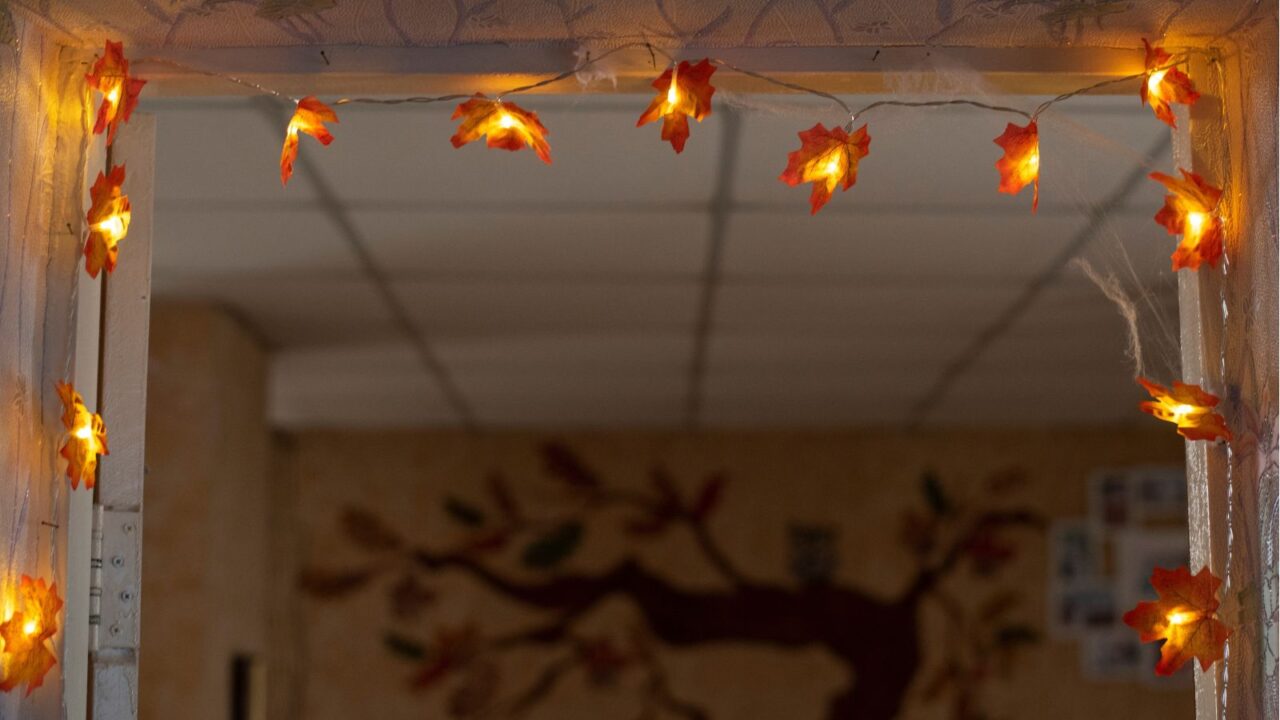 Fall interior decor. Orange electric lights having maple tree leaves shape attached to room door frame. Fall tree painting on the wall seen from door.