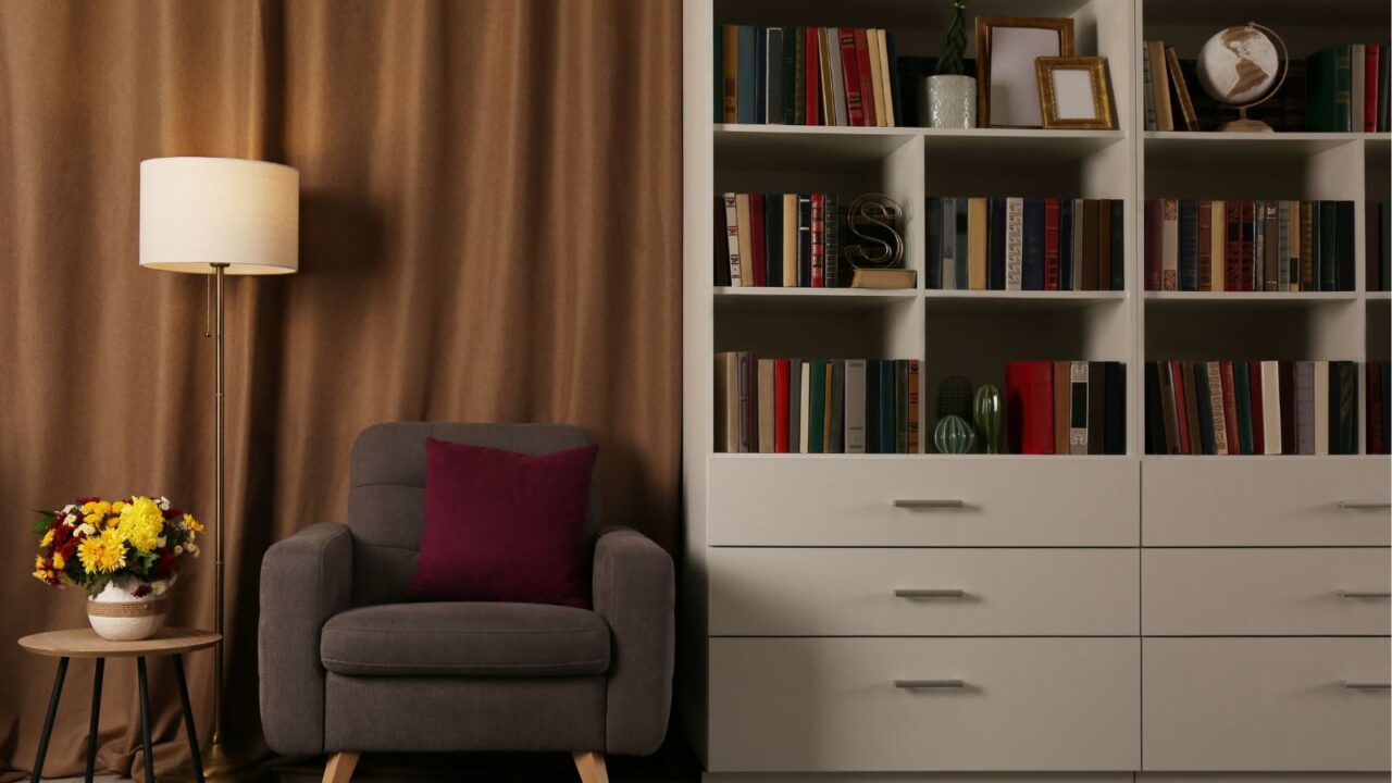 Cozy home library interior with comfortable armchair, floor lamp, a collection of books on a white bookshelf, and a wooden table with flowers in a vase.