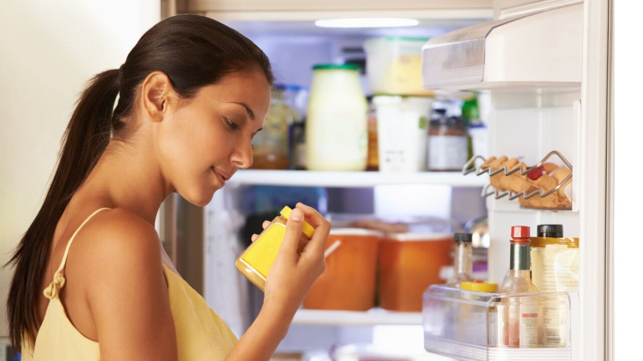 Open fridge and woman with sauce in hand checking label for preparing meal.