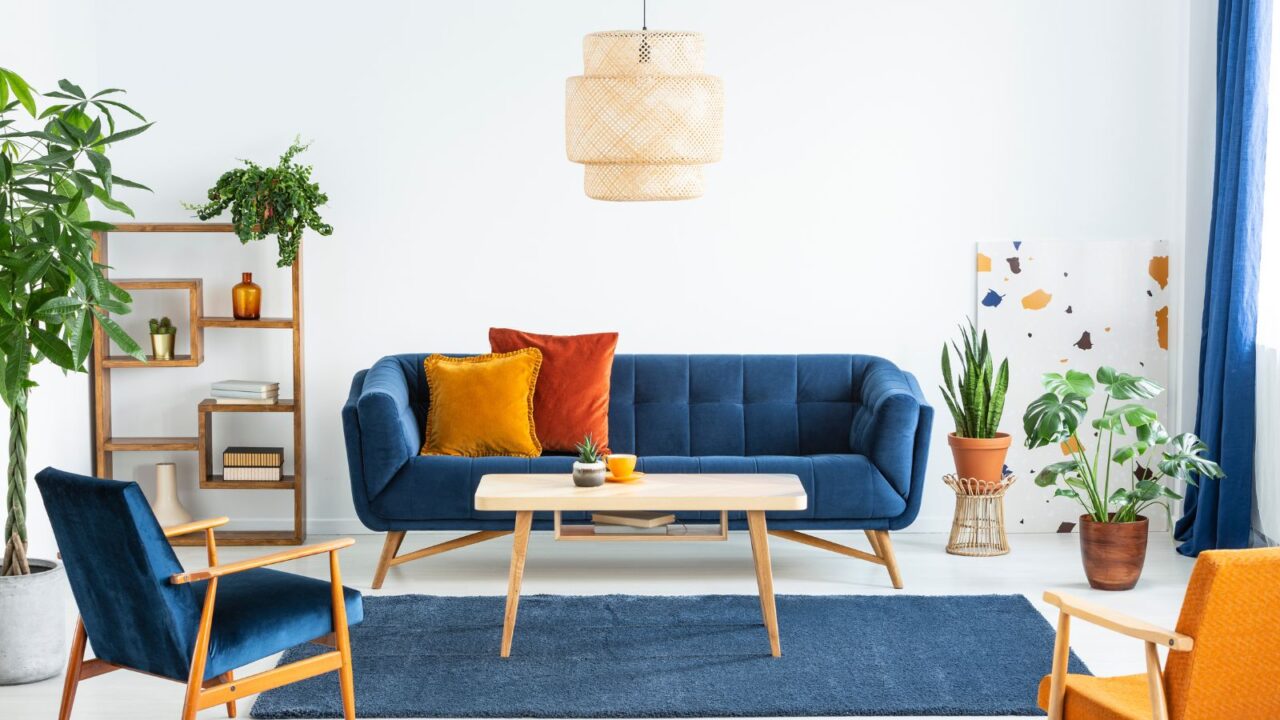 Retro inspired living room. Armchairs with wooden frame and colorful pillows on a navy blue sofa in a vibrant living room interior with green plants. A rattan light is hanging.