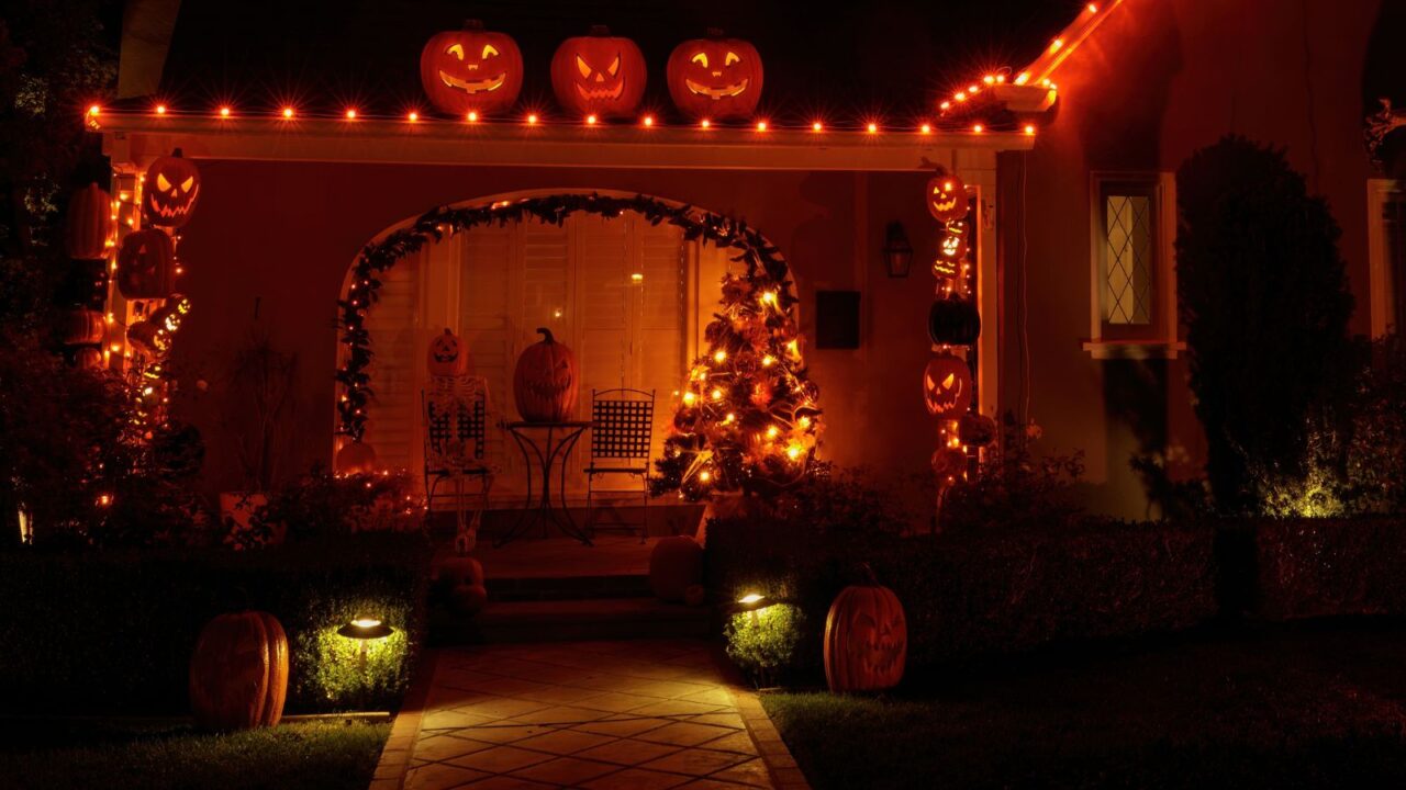 A Halloween decorated house with orange string lights, jack o lanterns, and carved pumpkins with a skeleton sitting on a chair on a porch.