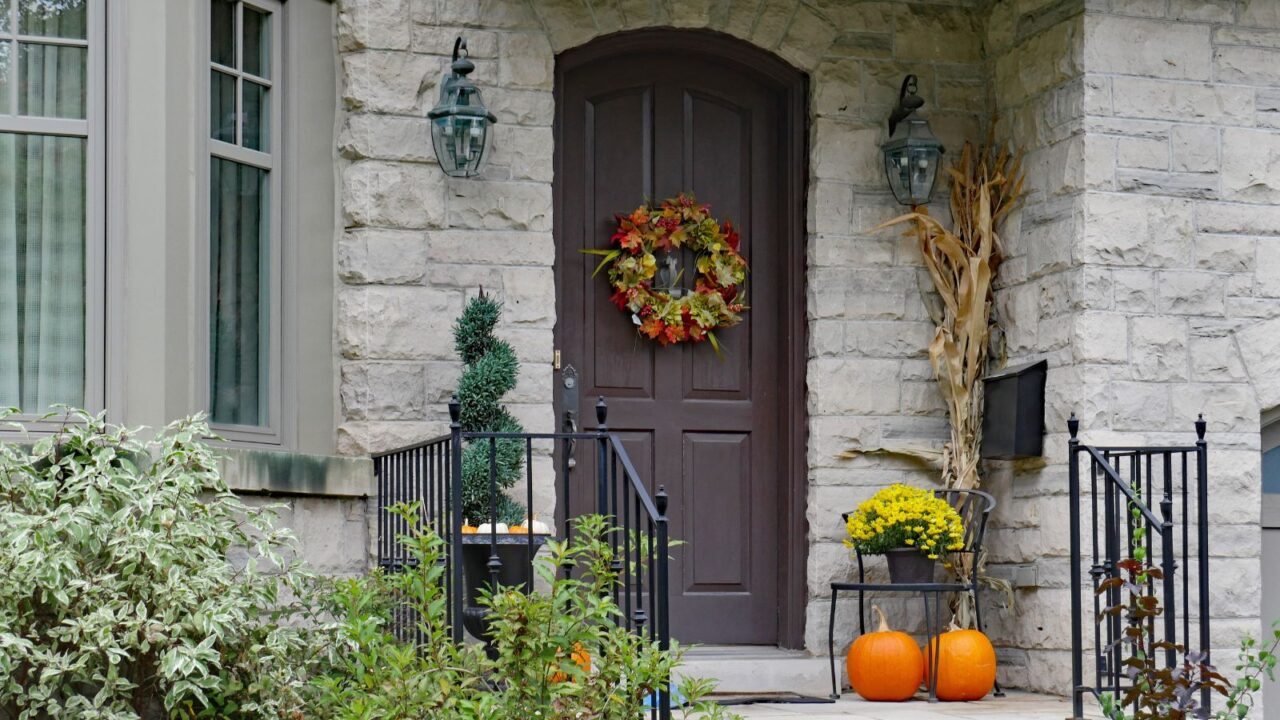 A colorful wreath on a brown front door of a house with stone walls with potted flowers, pumpkins, and dried corn stalk beside it.