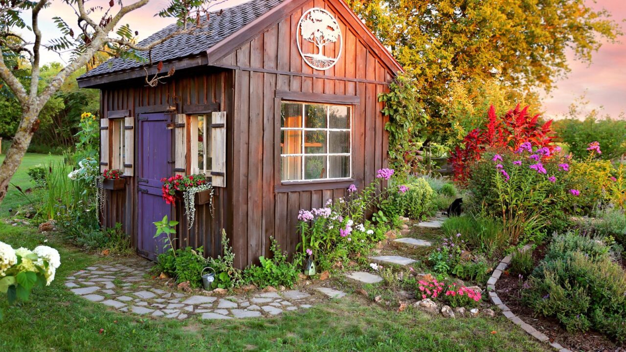 A wooden garden shed surrounded by bright flowers and plants, with a stone path.