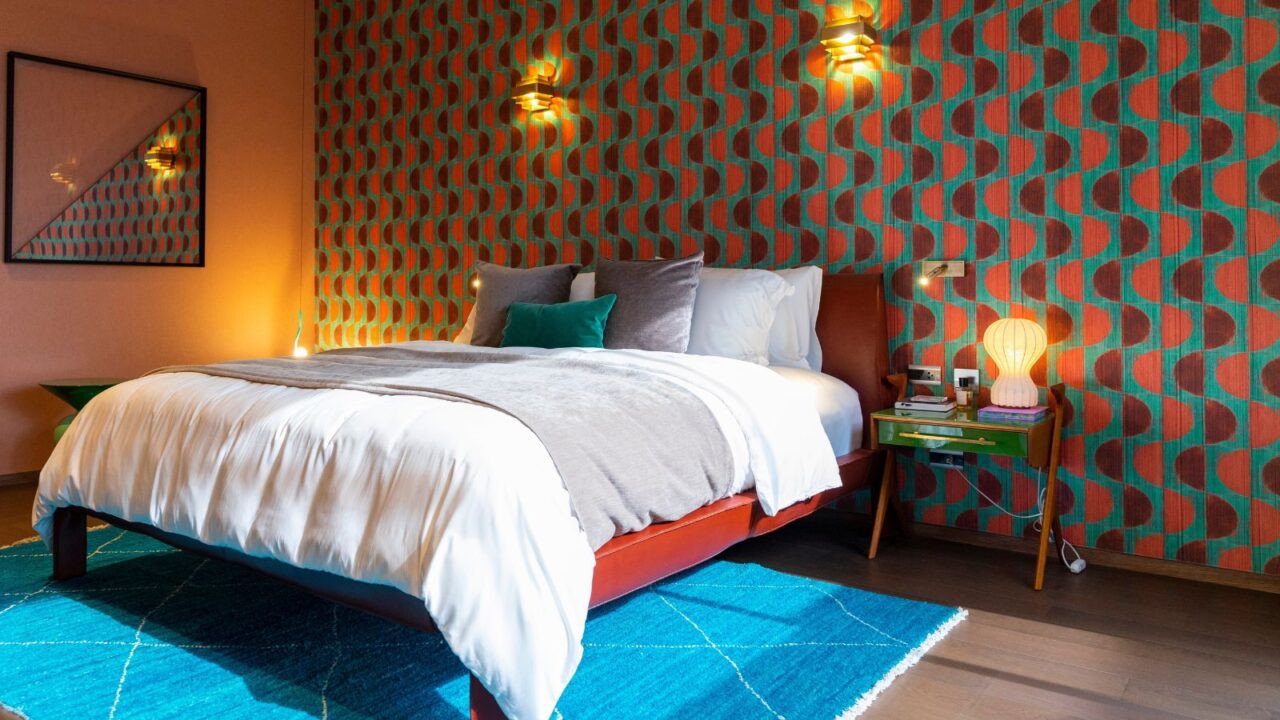 A bedroom interior with wooden bed with cushions, retro wallpaper, blue rug, a mirror, and a wooden side table with lamp and accessories.