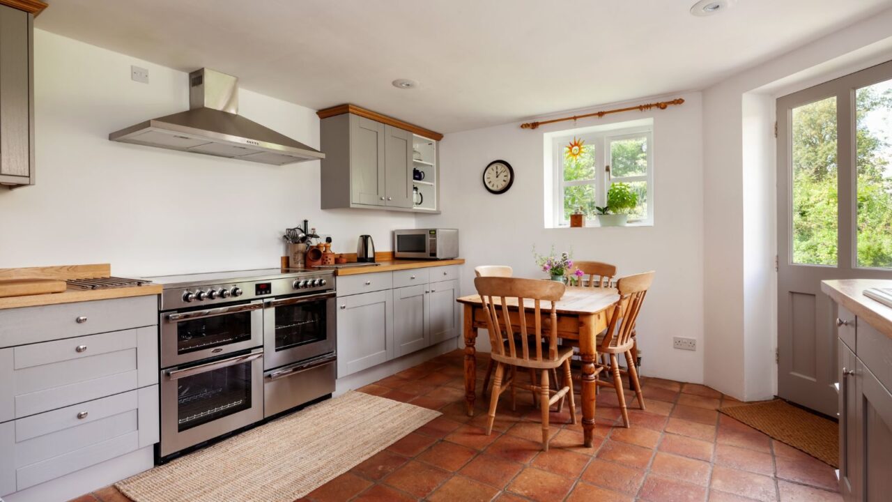 A cozy kitchen interior with terracotta flooring tiles, wooden dining table and chairs, and wooden cabinets.