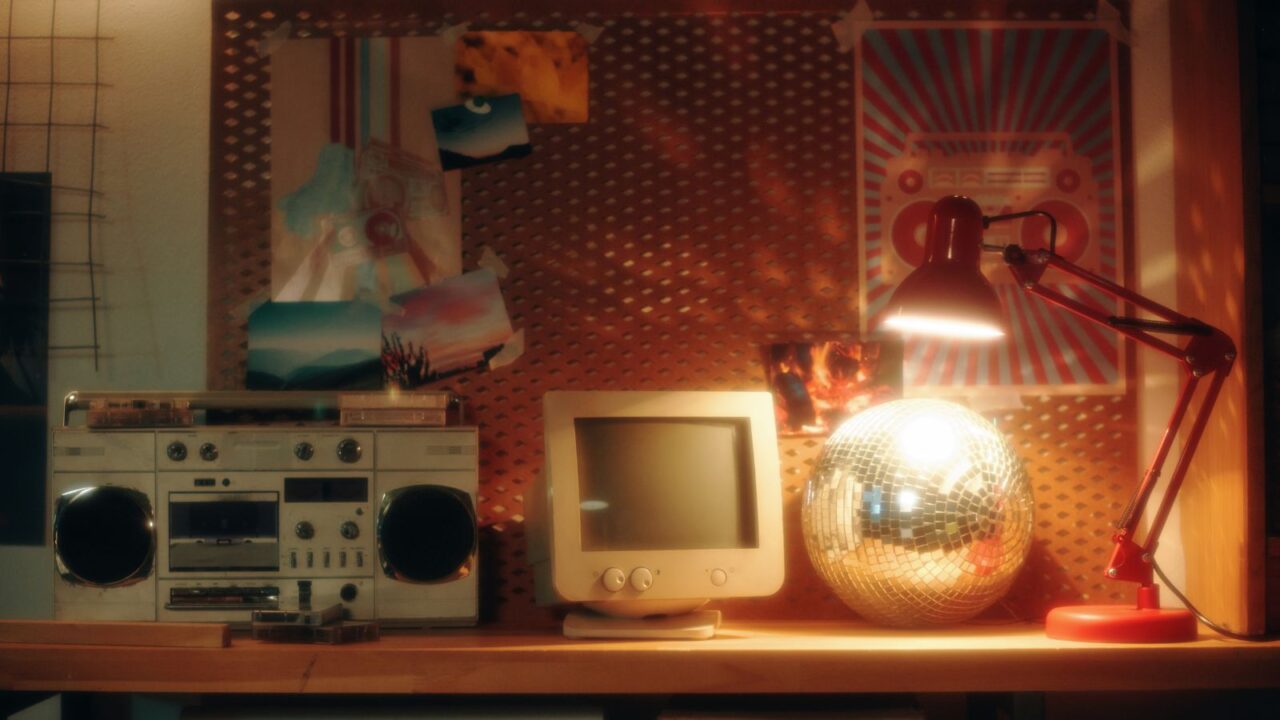 A retro room interior with a wooden table with boombox, lamp, an old desktop, and a pegboard with poster and polaroids