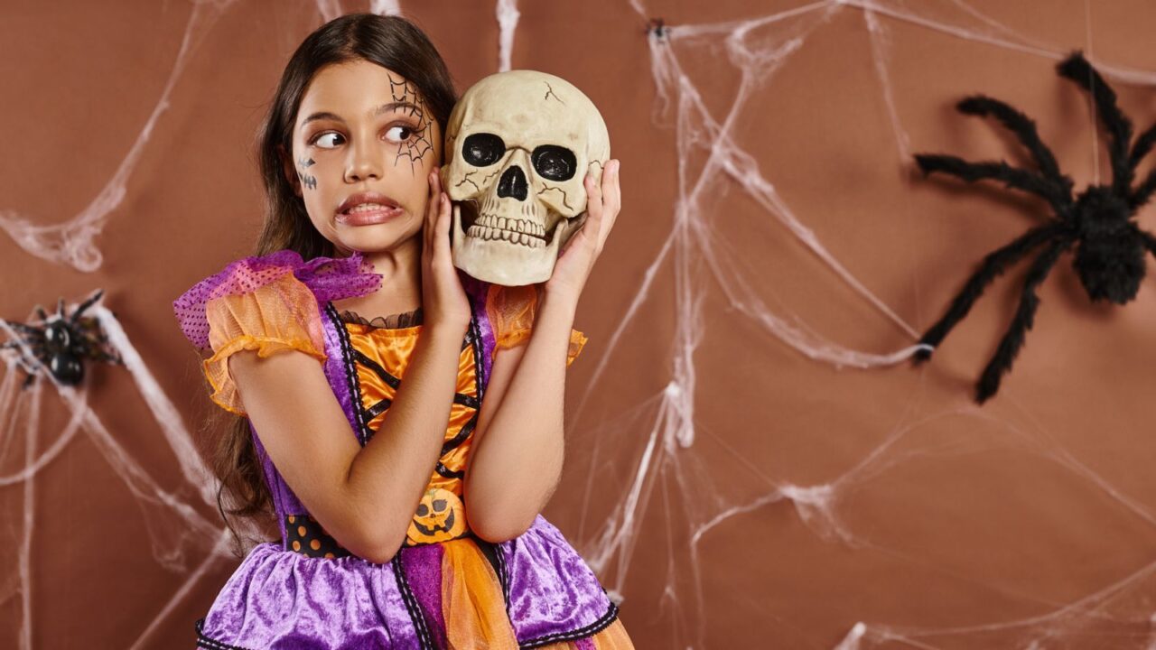 A girl in Halloween attire standing with skull and grimacing on brown background, spooky season.