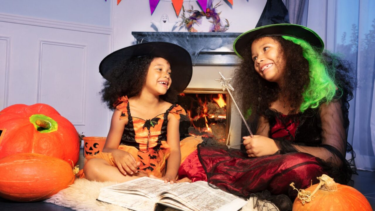 Two girls smiling and reading wizard book in Halloween witch costumes cast a spell with magic wand by fireplace.