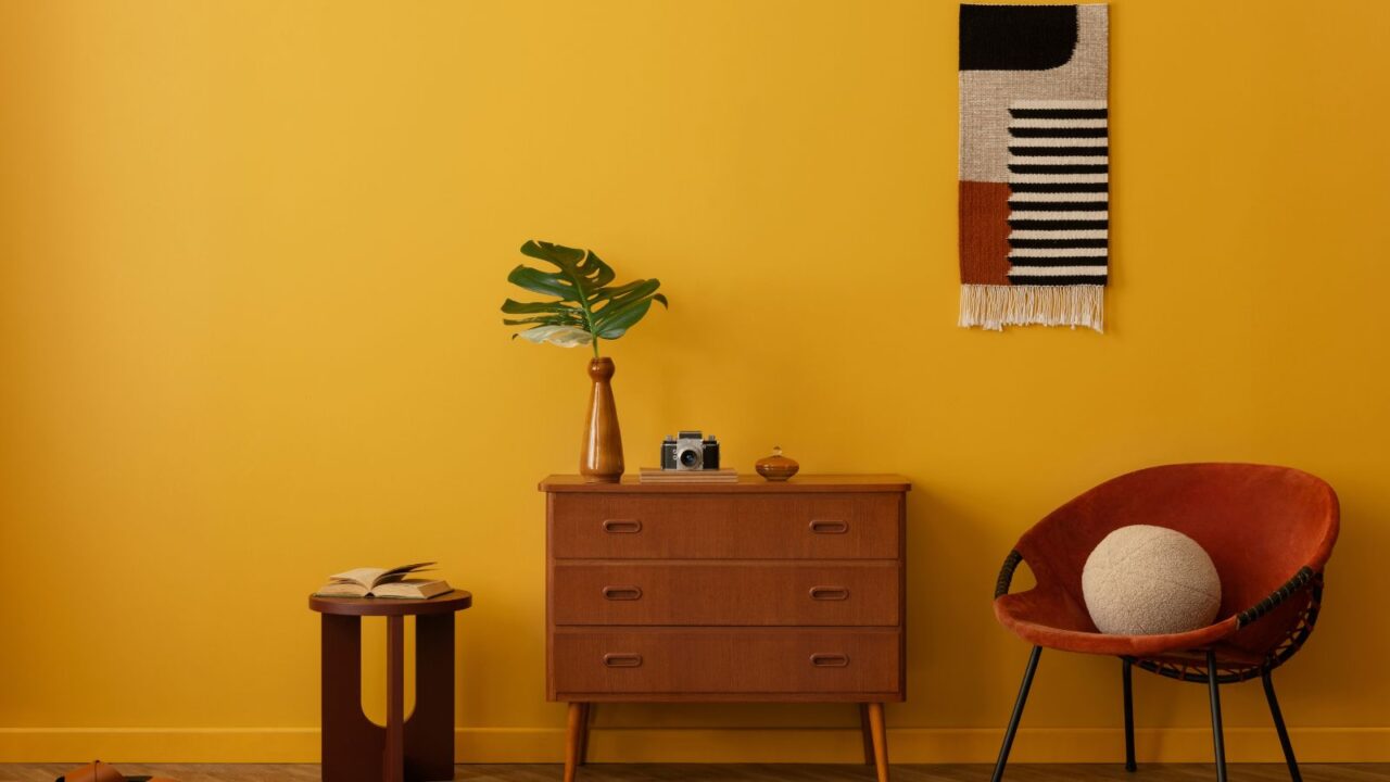 Minimalist vintage living room interior with red armchair, stylish wooden sideboard, burgundy stool, and yellow wall, and a wall hanging. Home decor.