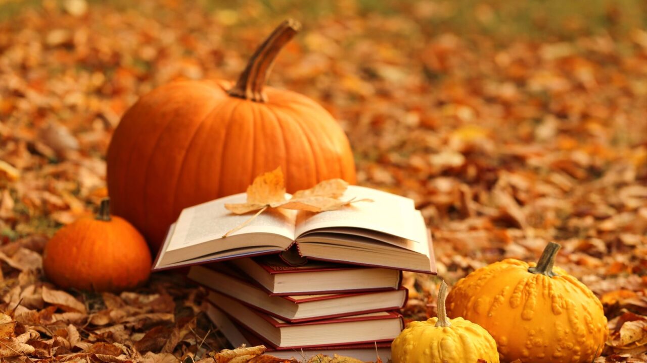 Reading fall books. Stack of books and orange pumpkins set on autumn foliage on blurred background.