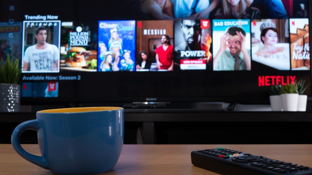 Close up of a coffee mug and remote control on a table with Netflix running on the television in background